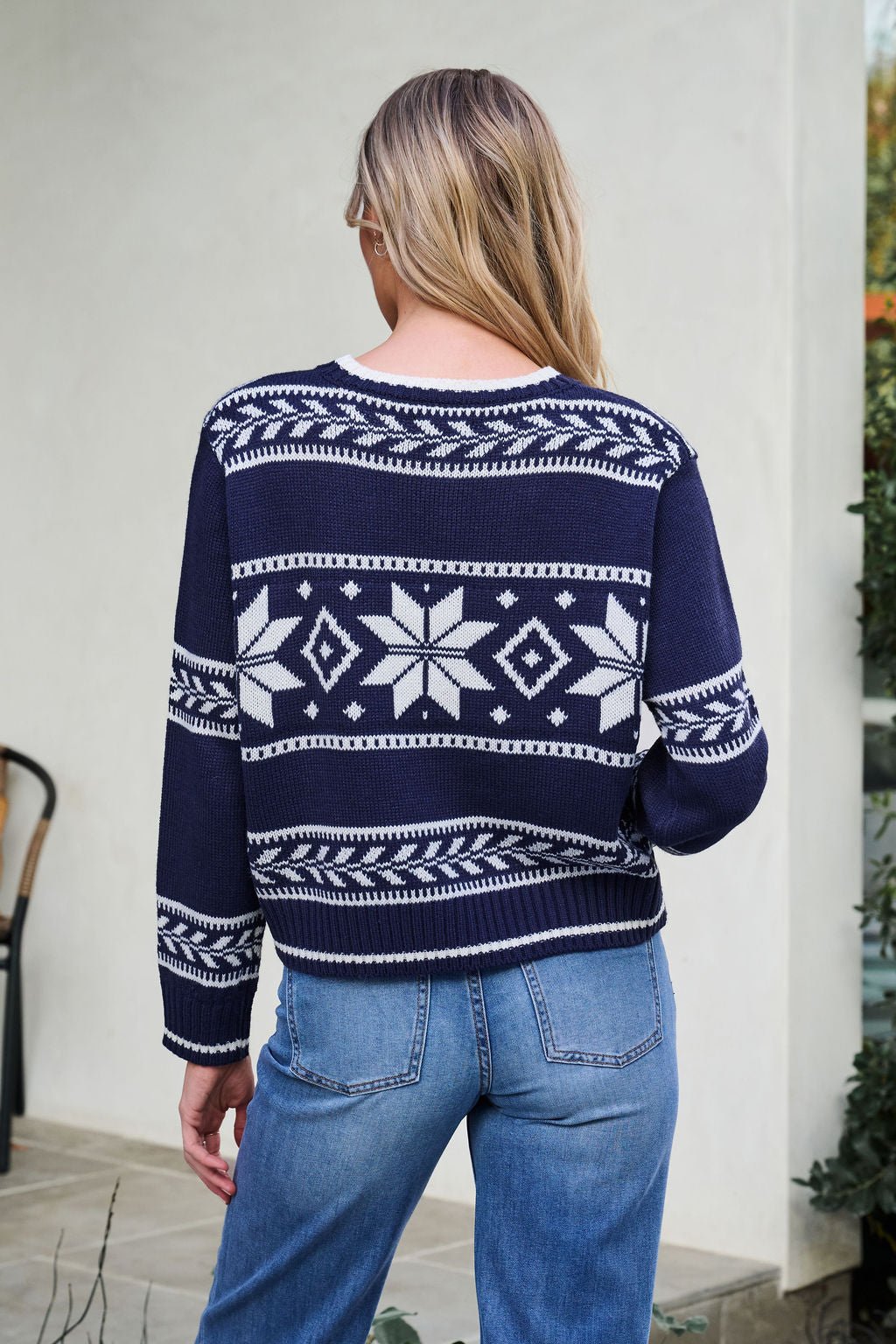 A woman with long blonde hair wears a Navy Ribbed Winter Snowflake Sweater and blue jeans, facing away from the camera.