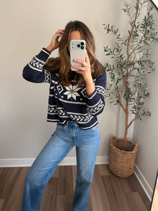 Woman wearing a Navy Ribbed Winter Snowflake Sweater and jeans takes a mirror selfie near a plant in a woven basket.