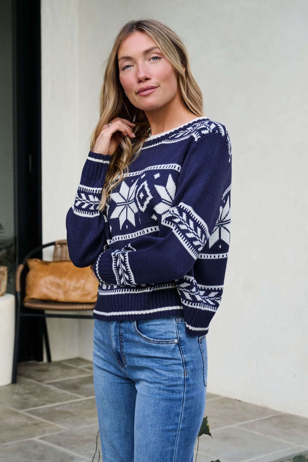 Woman with long blonde hair wears the Navy Ribbed Winter Snowflake Sweater and blue jeans, standing indoors by a chair and plant.