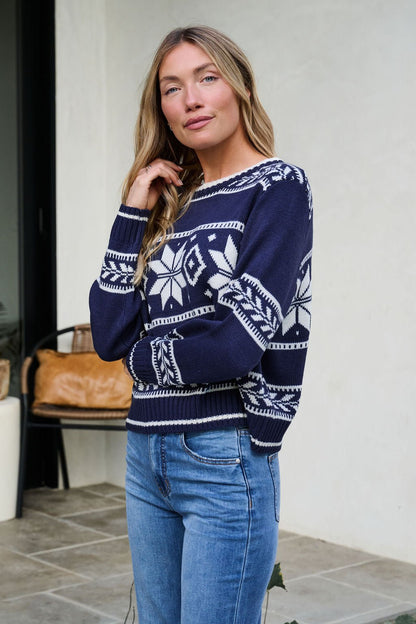 Woman with long blonde hair wears the Navy Ribbed Winter Snowflake Sweater and blue jeans, standing indoors by a chair and plant.