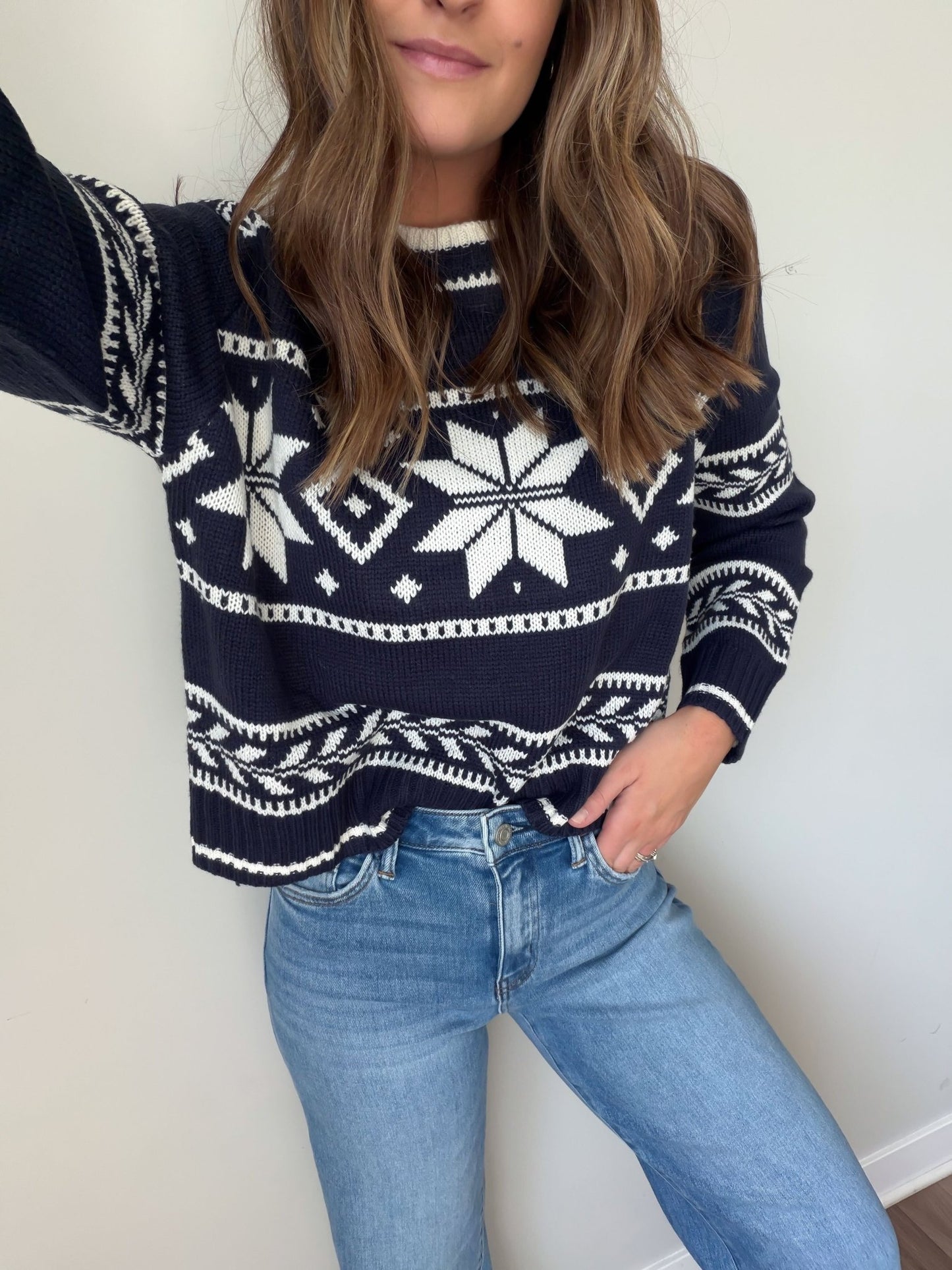 Woman in a Navy Ribbed Winter Snowflake Sweater and light blue jeans, standing against a plain light-colored wall.