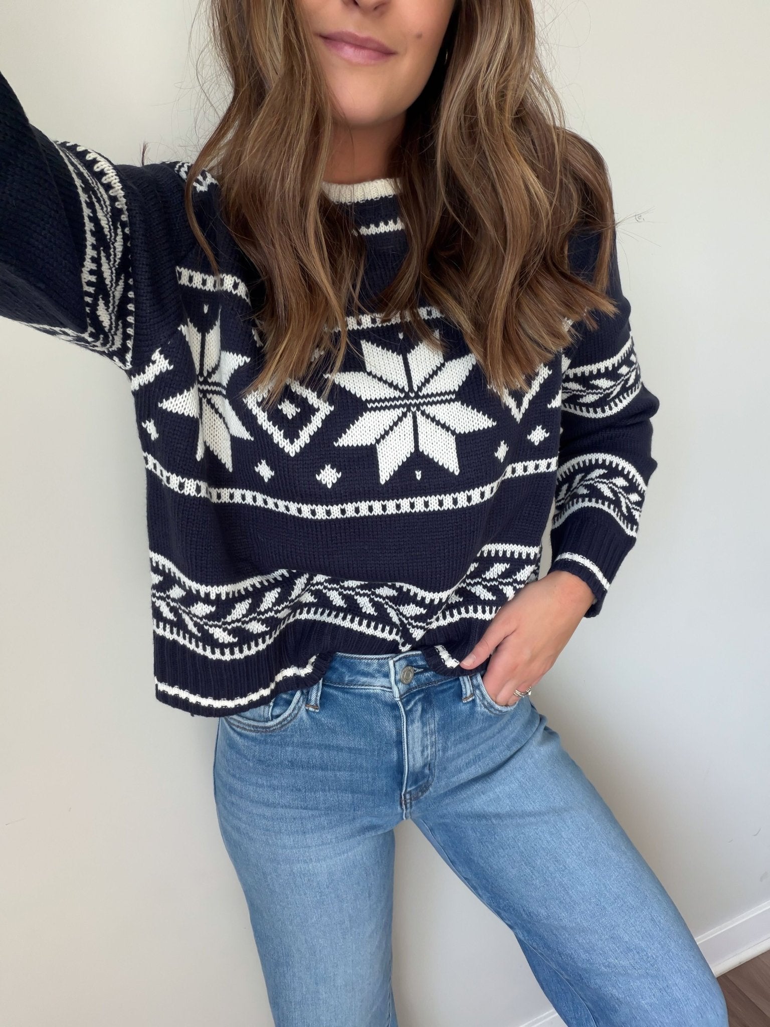 Woman in a Navy Ribbed Winter Snowflake Sweater and light blue jeans, standing against a plain light-colored wall.