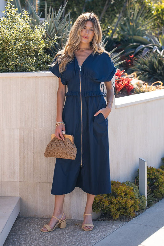 Wearing a Navy Smocked Zip Midi Dress, a woman stands outdoors with a woven handbag and beige sandals.