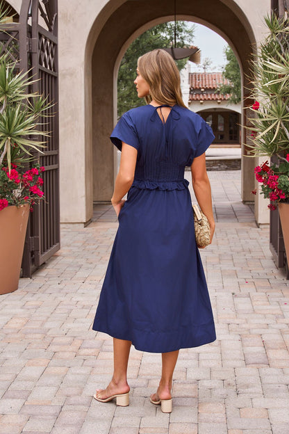 Woman in a Navy Smocked Zip Midi Dress and beige sandals holds a woven clutch outdoors, with flowers and arches around her.