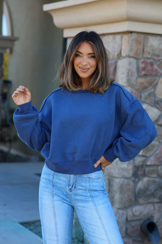 Woman with shoulder-length brown hair smiles outdoors by a stone wall, wearing a Navy Star Patch Sleeve Pullover and light-wash jeans.