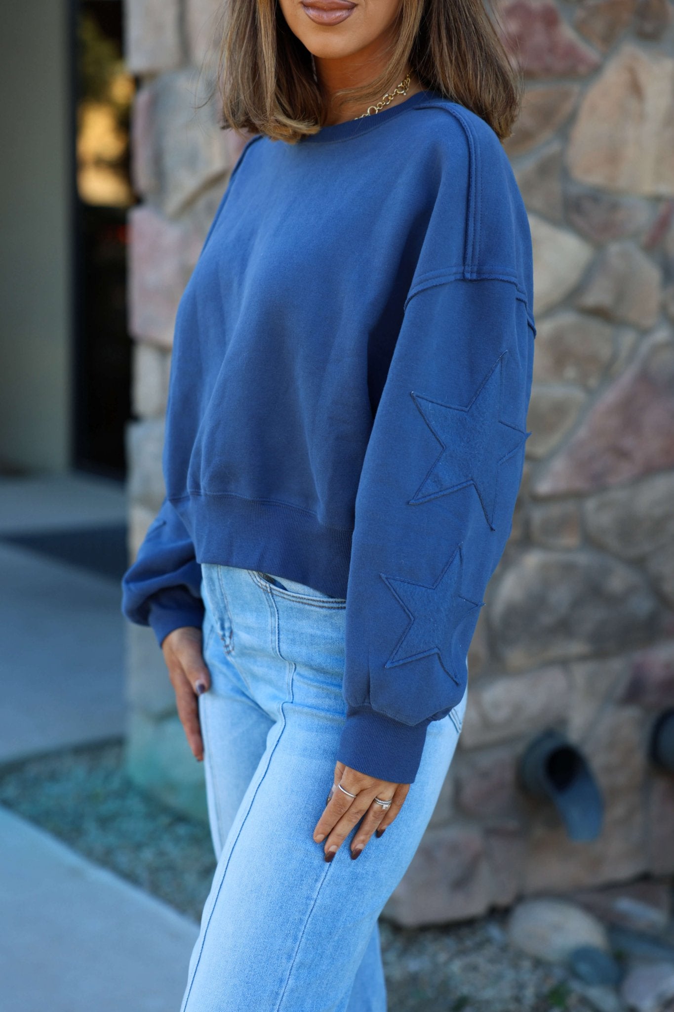 Woman wears a Navy Star Patch Sleeve Pullover and light blue jeans, standing outdoors by a stone wall.