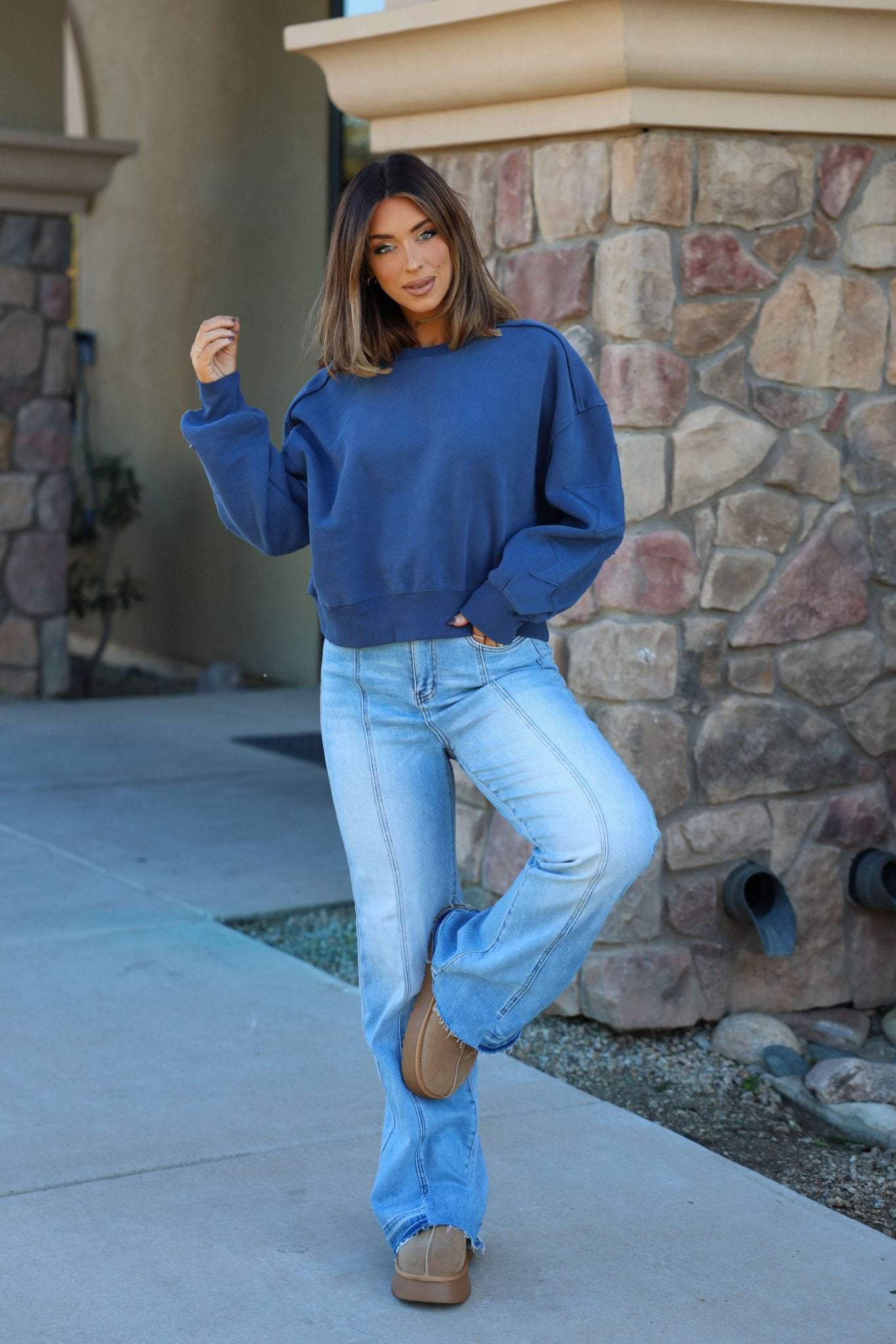Woman in a Navy Star Patch Sleeve Pullover and light jeans poses on one leg near a stone building, wearing tan platform shoes.