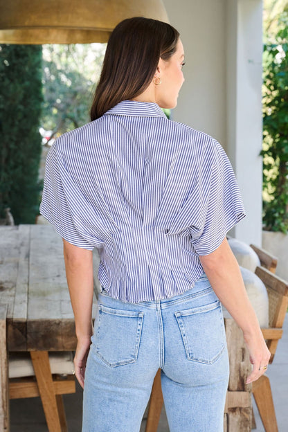A woman wearing a Navy Stripe Pleated Button Down Shirt and light blue jeans stands outdoors near a wooden table, back to the camera.