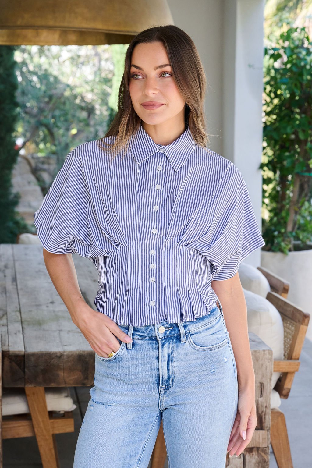 A woman wears the Navy Stripe Pleated Button Down Shirt with light-wash jeans, standing indoors and smiling with one hand in her pocket.