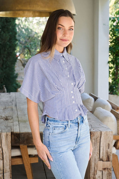 A woman in a Navy Stripe Pleated Button Down Shirt and light jeans stands by a rustic wooden table, smiling gently at the camera.