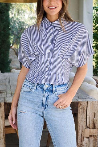 Woman wearing a Navy Stripe Pleated Button Down Shirt and high-waisted light blue jeans, standing by a rustic table.
