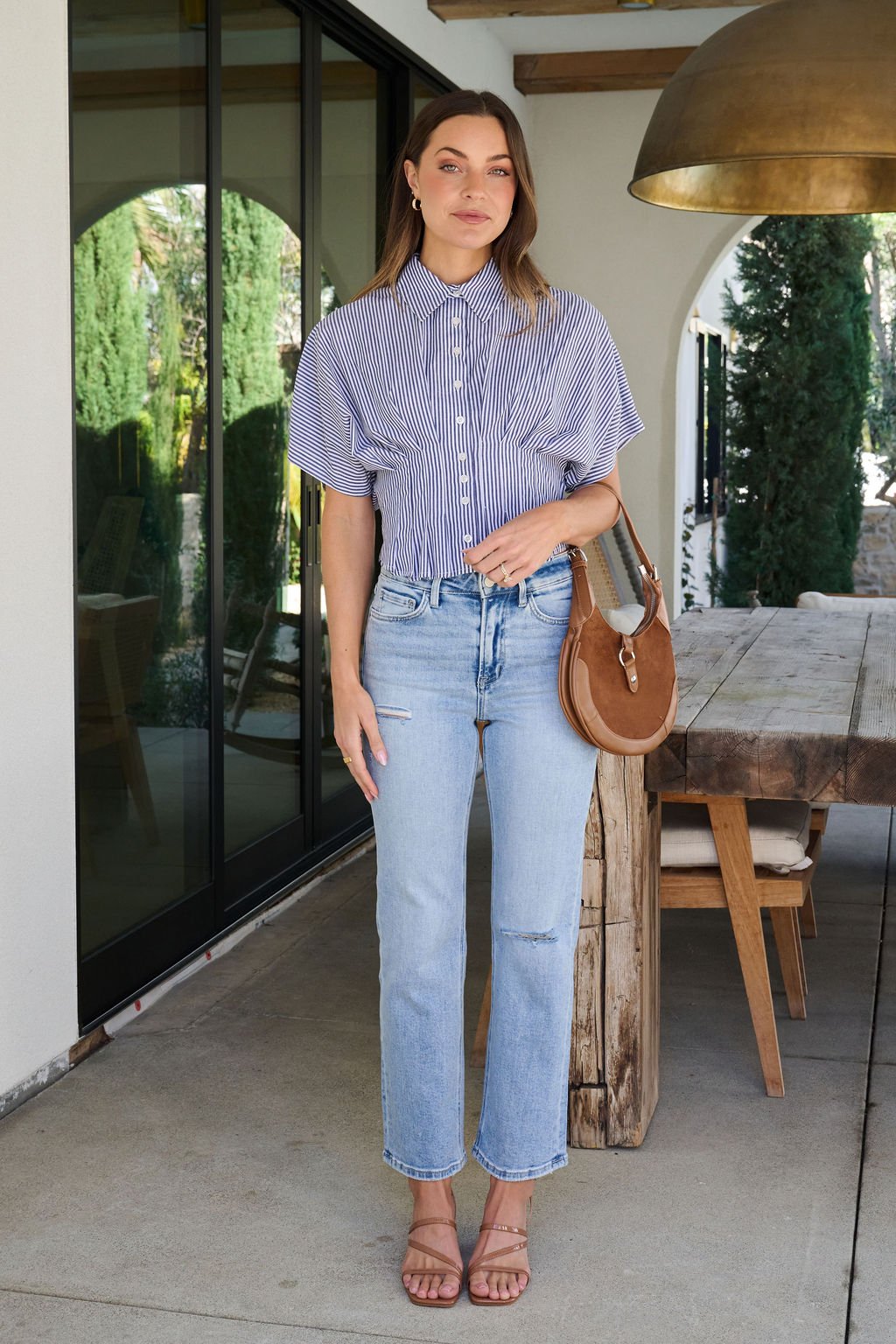 A woman stands outside by a wooden table, wearing the Navy Stripe Pleated Button Down Shirt, light blue jeans, sandals, and a brown bag.
