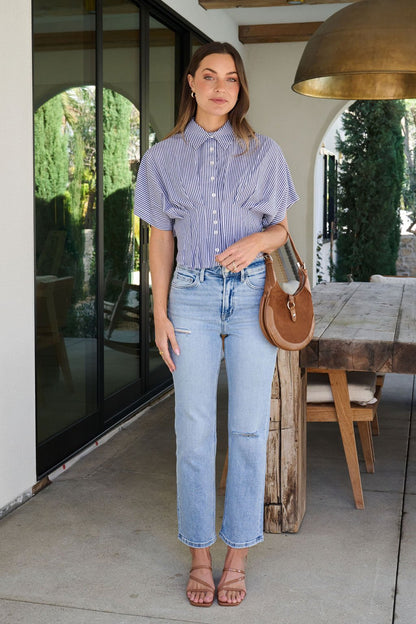 A woman stands outside by a wooden table, wearing the Navy Stripe Pleated Button Down Shirt, light blue jeans, sandals, and a brown bag.