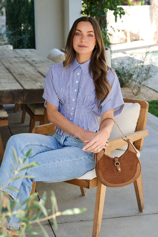 A woman sits outside on a wooden chair, wearing the Navy Stripe Pleated Button Down Shirt with jeans and a brown handbag.