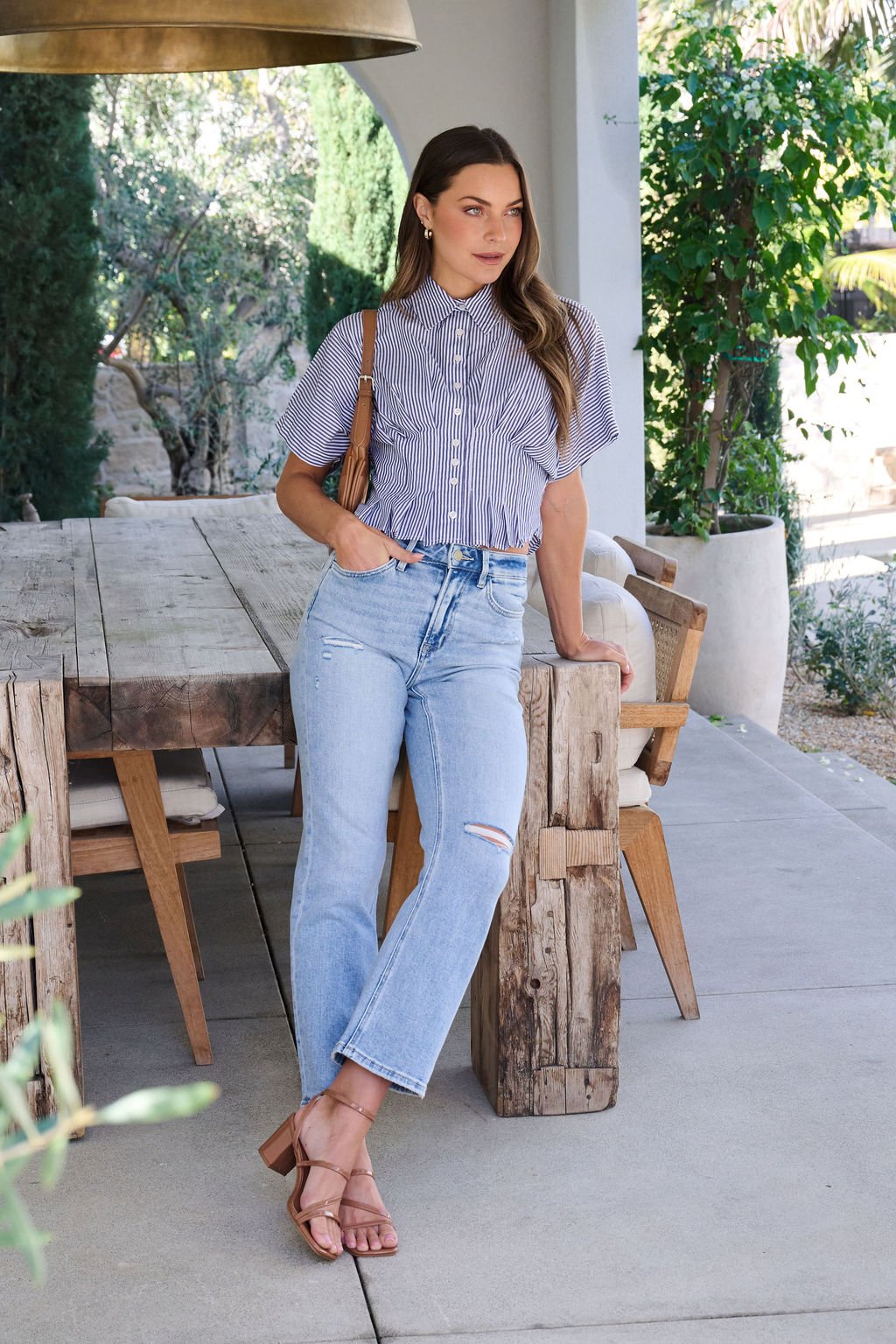 A woman in a Navy Stripe Pleated Button Down Shirt and ripped jeans leans on an outdoor wooden table, holding a purse.