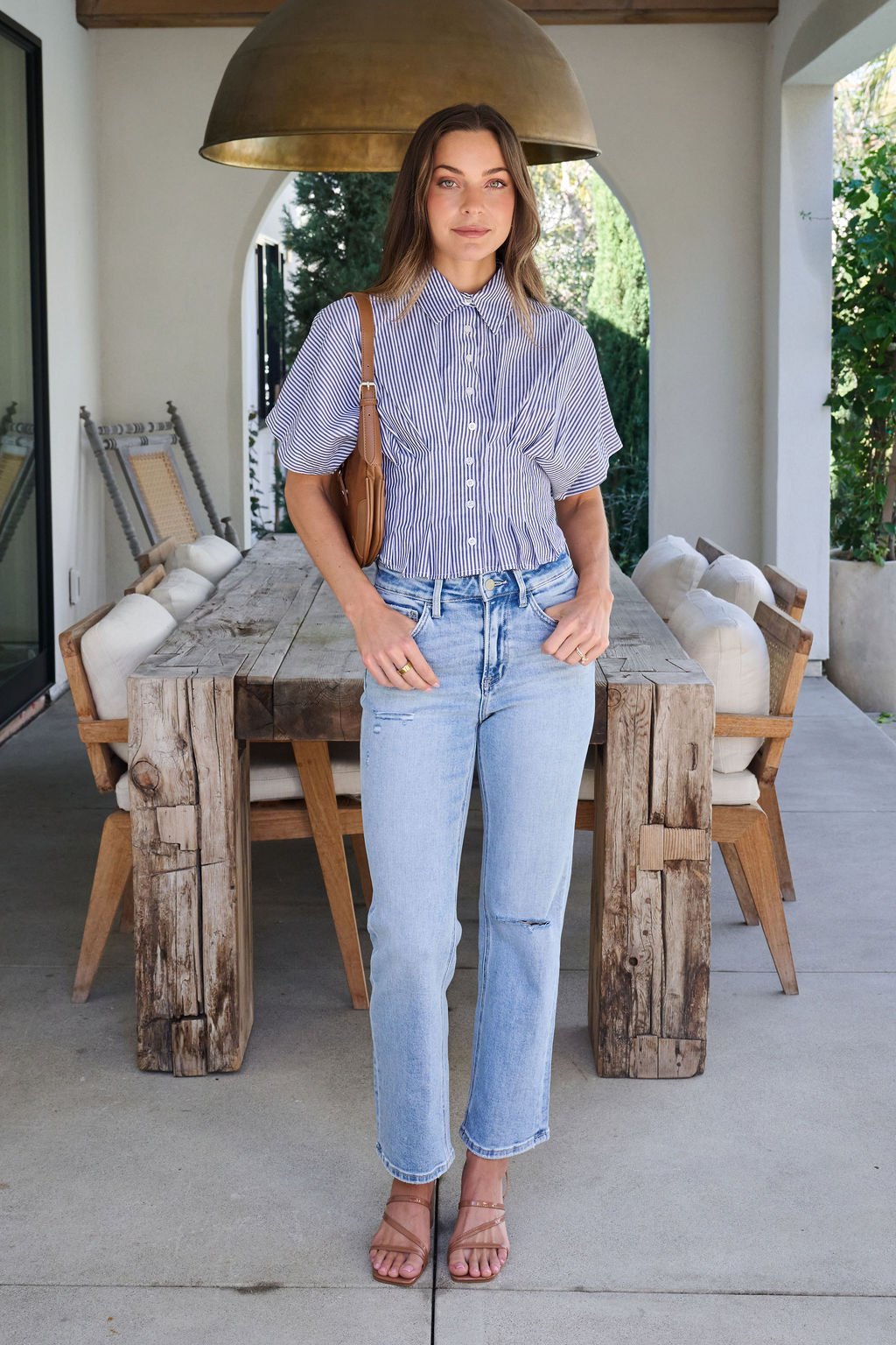 A woman outdoors by a rustic table wears a Navy Stripe Pleated Button Down Shirt, light jeans, sandals, and a brown shoulder bag.