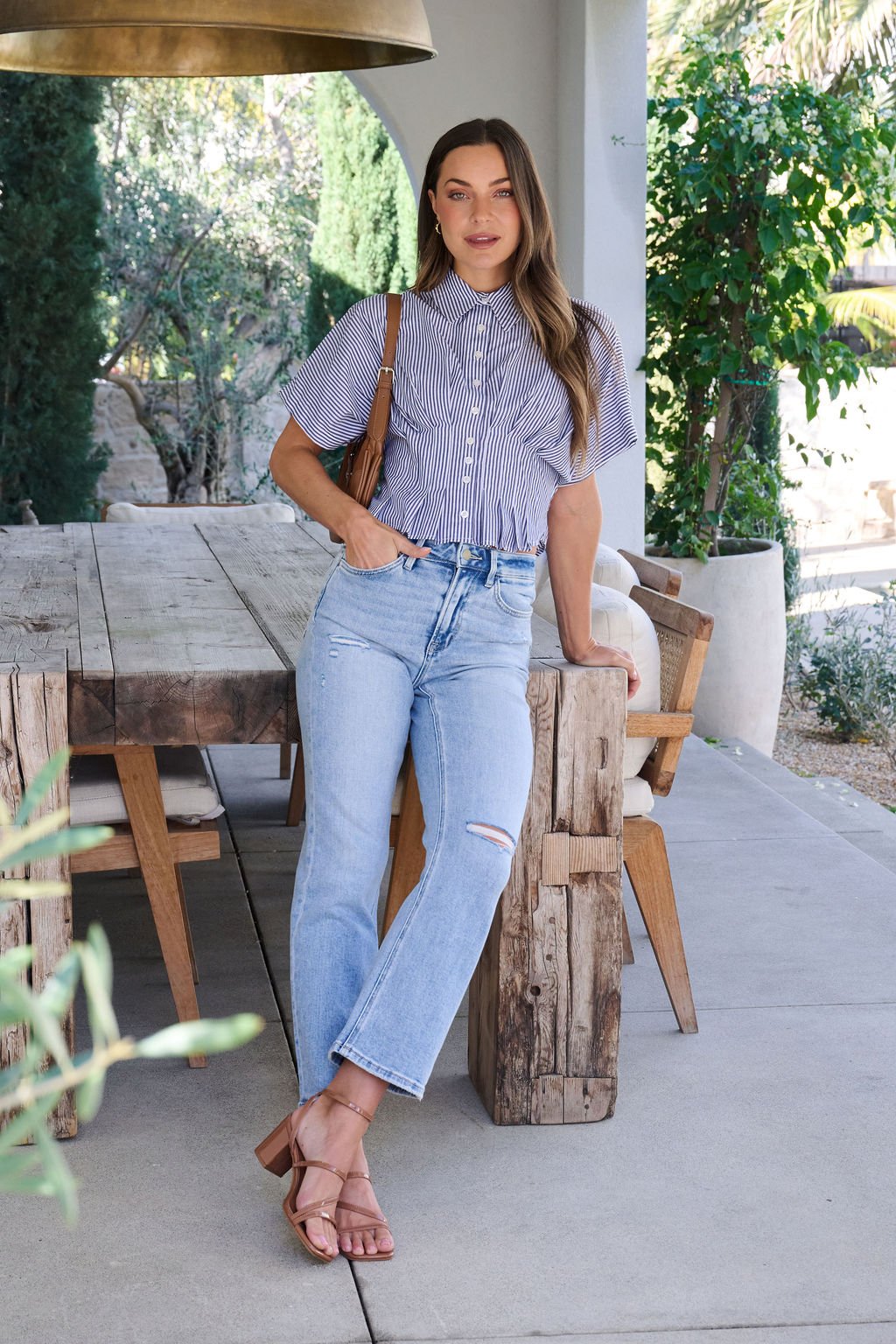 A woman in a Navy Stripe Pleated Button Down Shirt and ripped jeans leans on a rustic outdoor table amid greenery.
