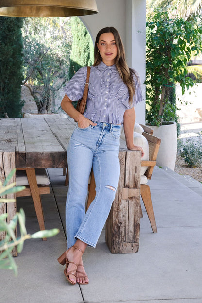 A woman in a Navy Stripe Pleated Button Down Shirt and ripped jeans leans on a rustic outdoor table amid greenery.