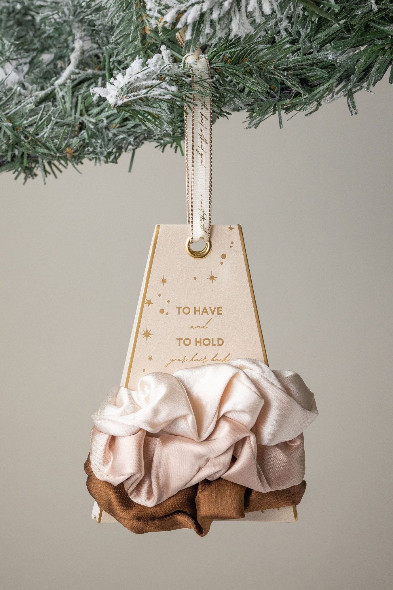 To Have and To Hold" gift tag hangs on a frosted pine with the Neutral Gaja Satin Scrunchie Set in cream, blush, and brown.
