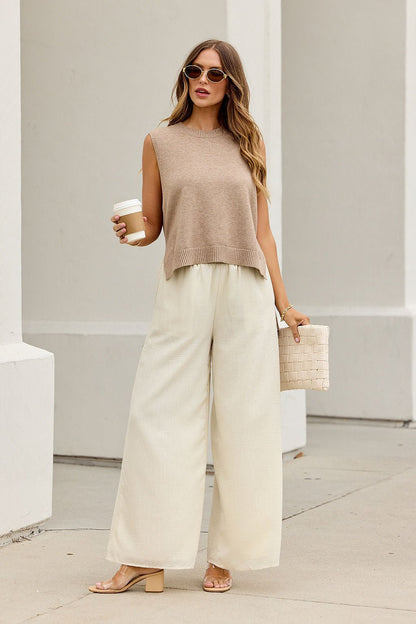 Woman in sunglasses wears the Nine To Five Cashmere Sweater in dark beige, wide cream pants, holding coffee and a clutch on a city sidewalk.