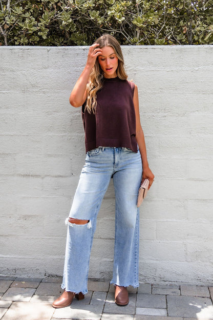 Woman in a Nine To Five Cashmere Sweater - Mocha and ripped jeans stands against a white wall, holding a beige clutch.