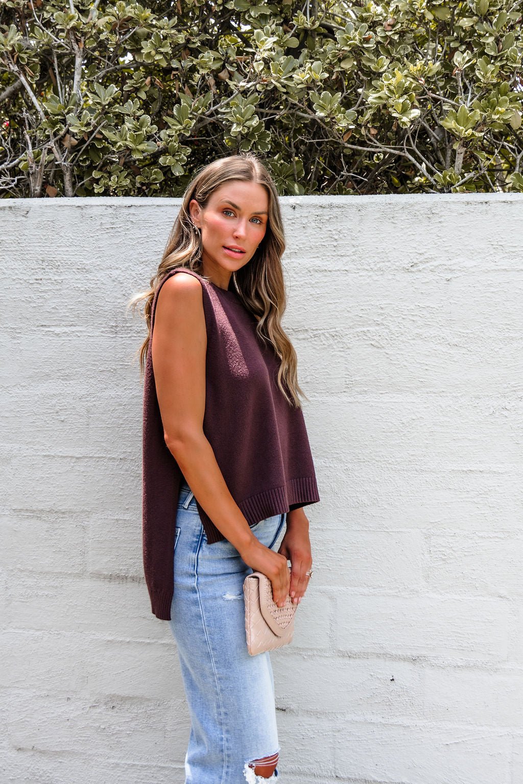 Woman in a Nine To Five Cashmere Sweater - Mocha and ripped jeans stands by a white wall, holding a beige clutch.