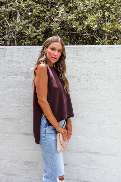Woman in a Nine To Five Cashmere Sweater - Mocha and ripped jeans stands by a white wall, holding a beige clutch.