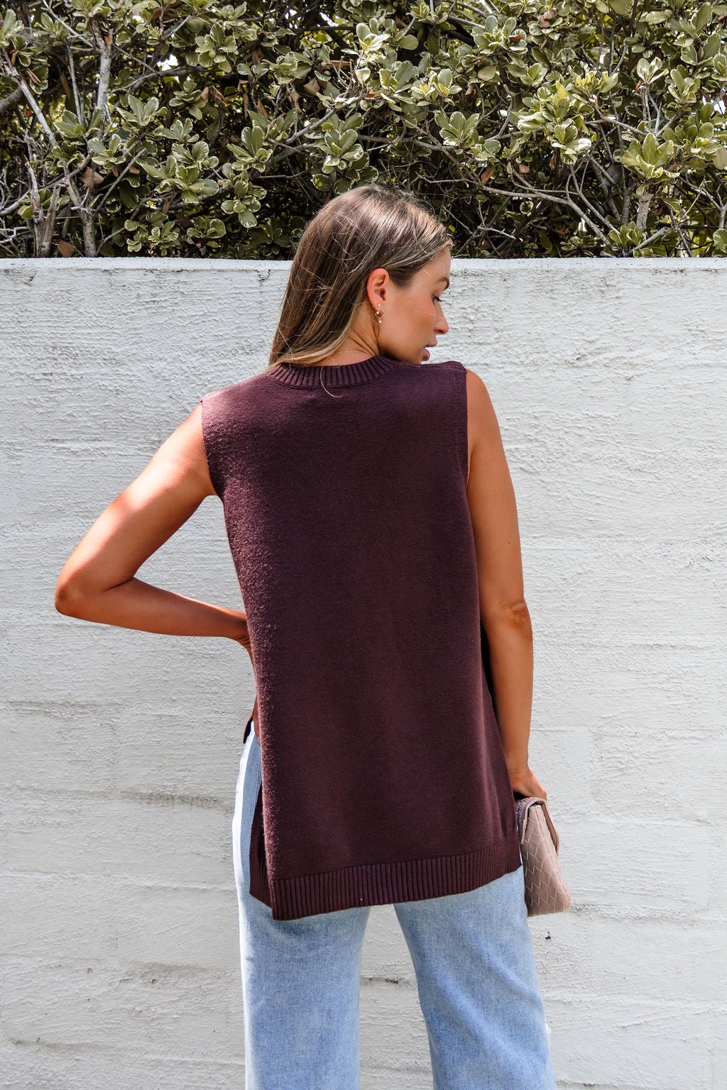 A woman in a relaxed fit Nine To Five Cashmere Sweater - Mocha and light jeans stands facing a white wall with plants above it.