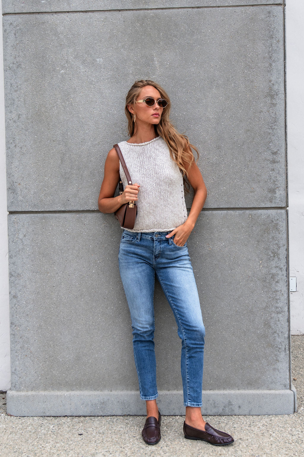 Woman in sunglasses, Free People Opal Tank Top, blue jeans, and loafers stands by a gray wall holding a brown shoulder bag.