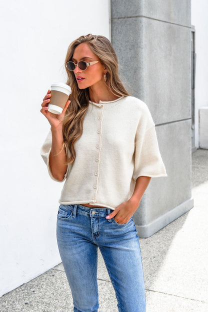 Woman in sunglasses wears the Corbin Button Down Sweater Cardigan in cream with jeans, holding a coffee cup by a stone wall.