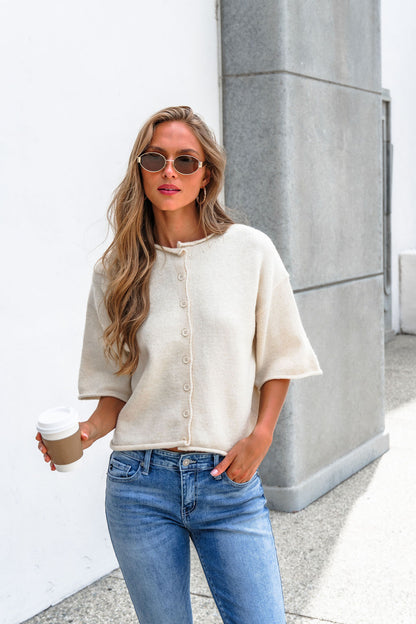 Woman in sunglasses wears Corbin Button Down Sweater Cardigan - Cream and jeans, holding coffee near a light gray building outdoors.