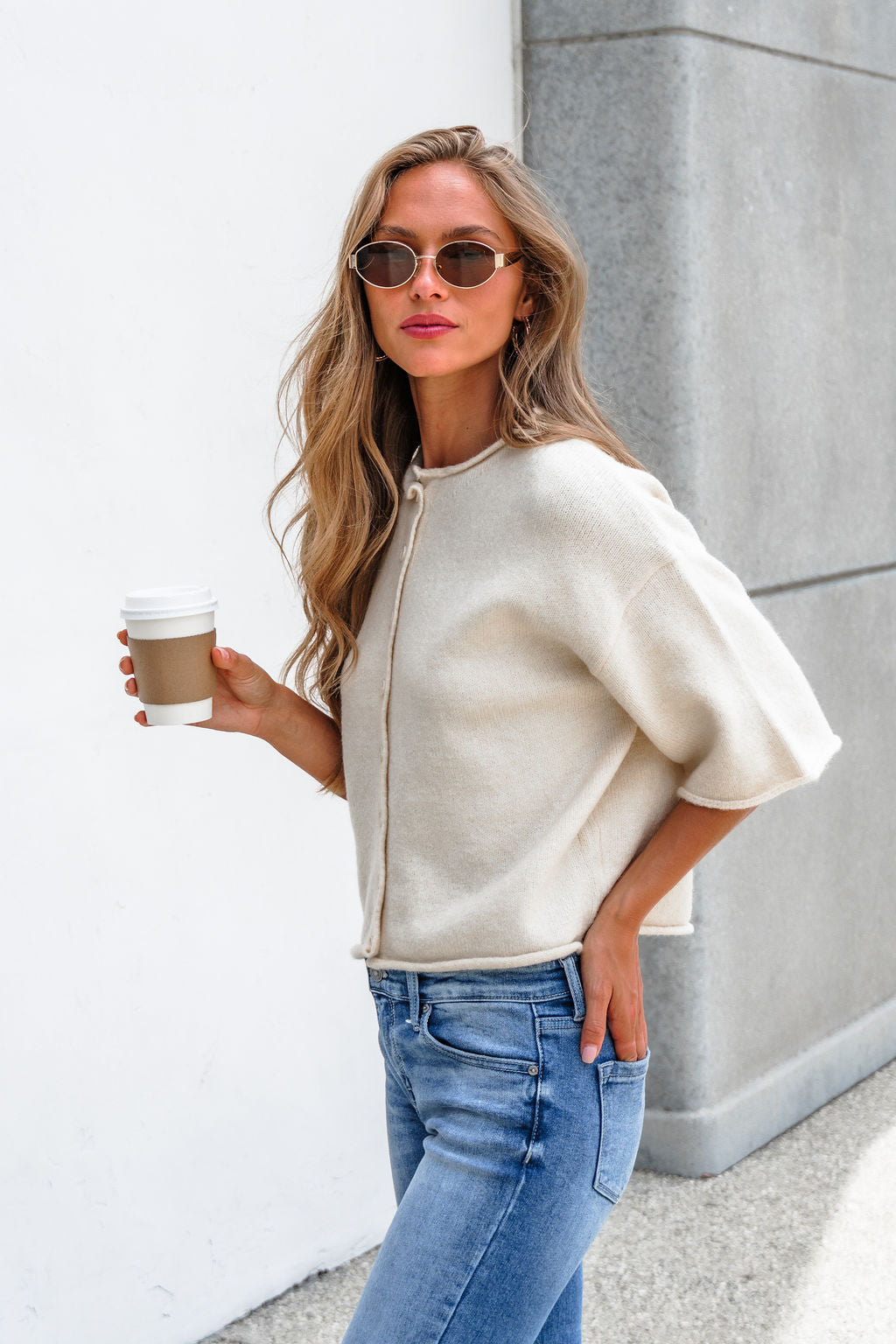 Woman in sunglasses wears the Corbin Button Down Sweater Cardigan - Cream, holding coffee, one hand in her jeans, by a gray wall.