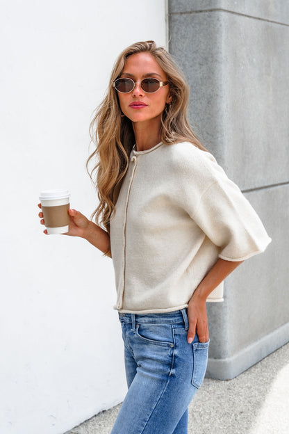 Woman in sunglasses wears the Corbin Button Down Sweater Cardigan - Cream, holding coffee, one hand in her jeans, by a gray wall.