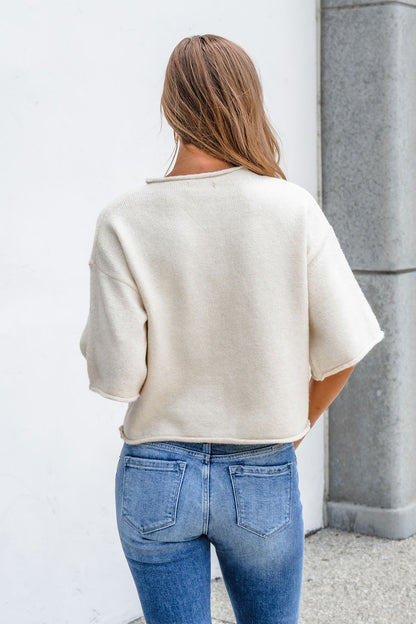 Woman with long hair wears the Corbin Button Down Sweater Cardigan - Cream and blue jeans, facing away near a white wall and gray pillar.