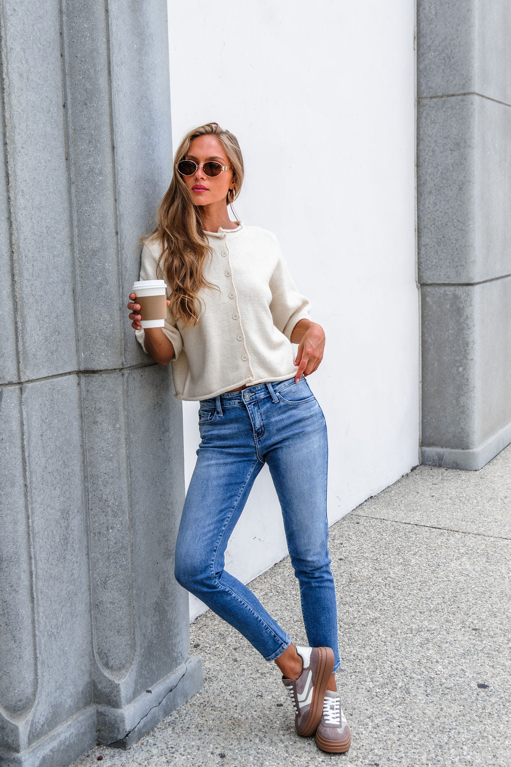 Woman in sunglasses and blue jeans leans against a gray wall wearing the Corbin Button Down Sweater Cardigan - Cream, holding a coffee cup.