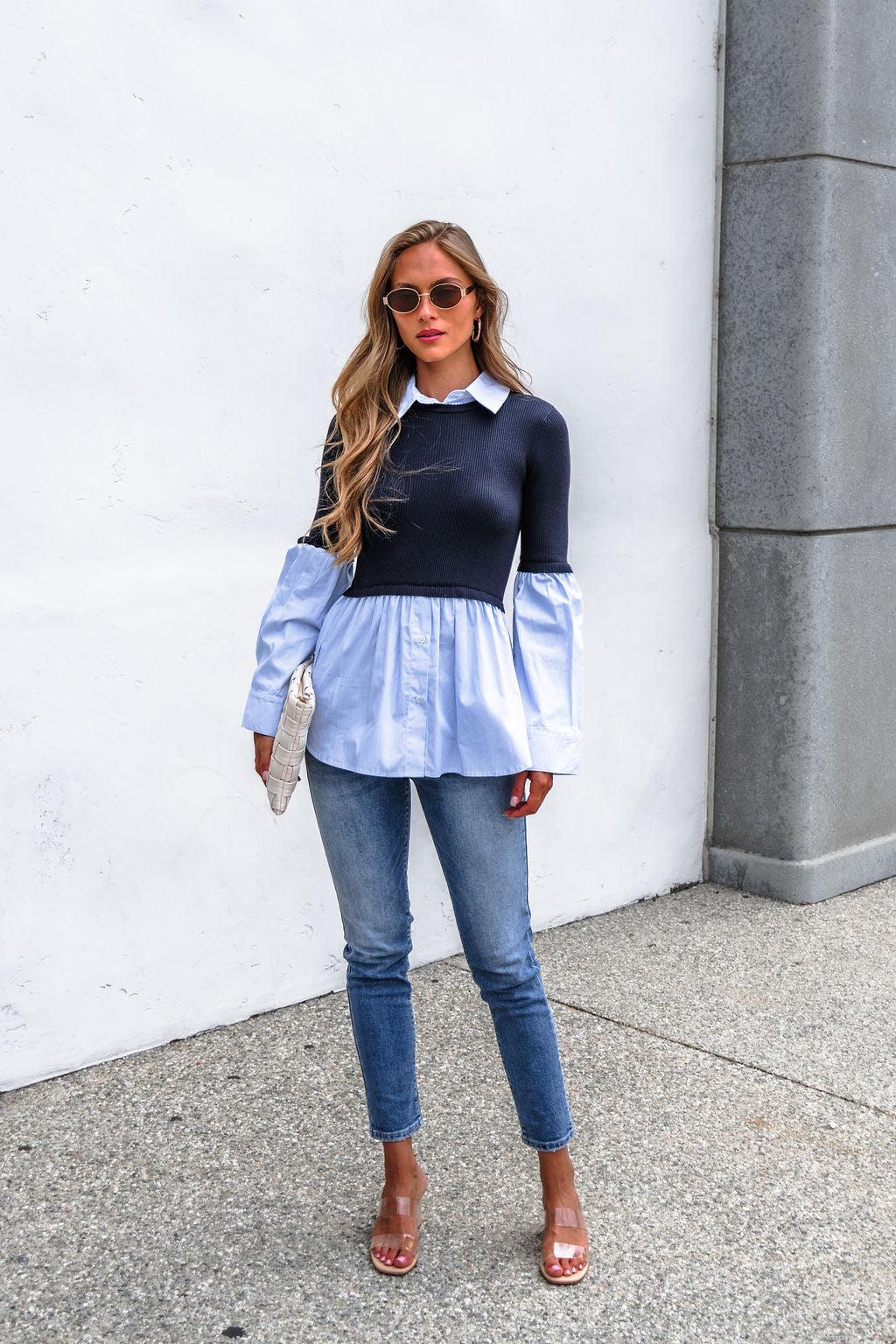 Woman in sunglasses, Steve Madden Lenny Top - Navy, jeans, clear heels, with a textured white clutch against a white wall.
