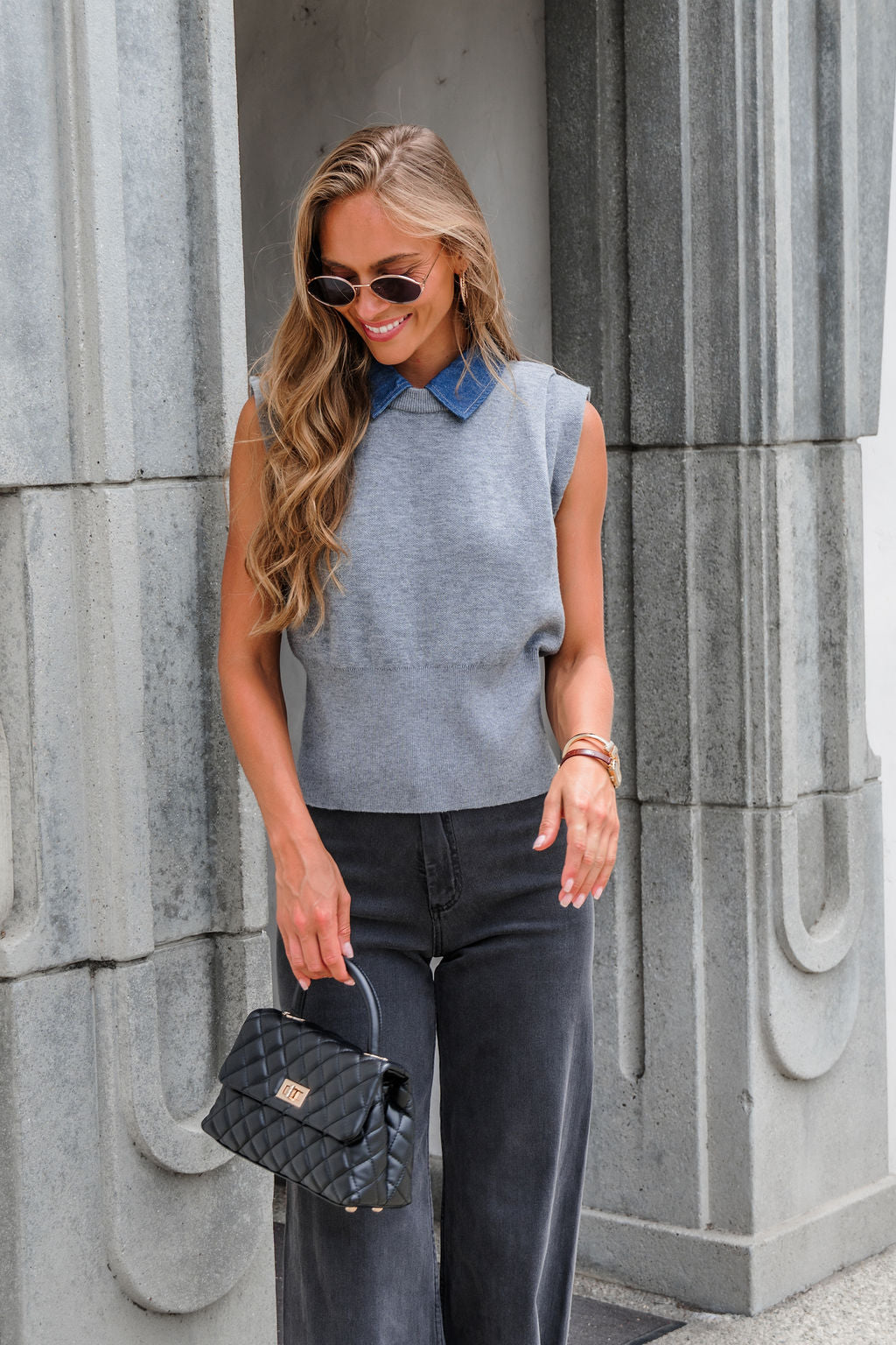 Woman in sunglasses and the Steve Madden Kelson Sweater - Grey, holding a black quilted handbag, stands by a gray stone wall.