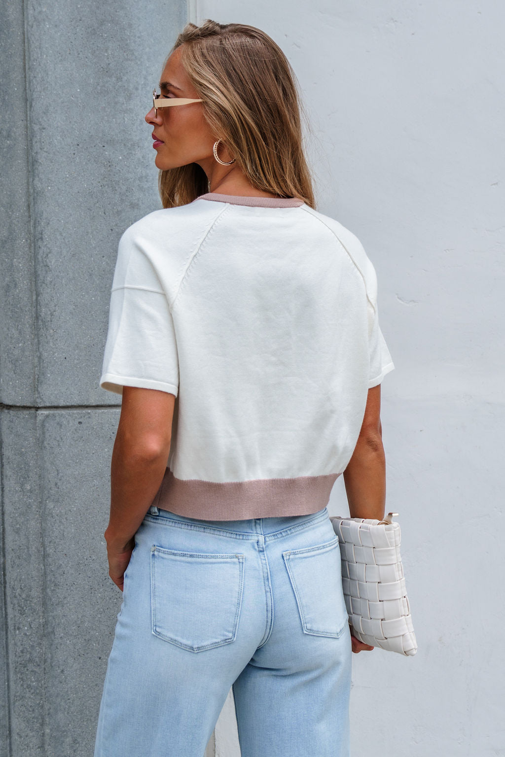 Woman with long hair in sunglasses wears the Contrast Button Front Sweater - Cream and light blue jeans, holding a white woven clutch.