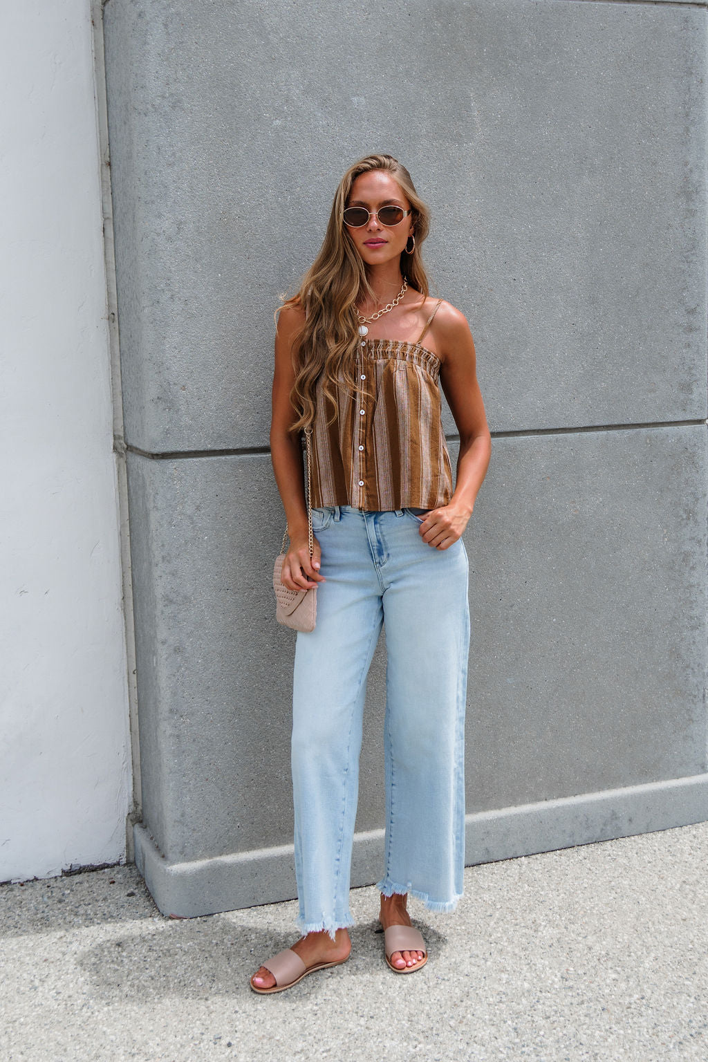 Woman in sunglasses wears the Sadie & Sage Striped Linen Cami - Camel, light blue jeans, and sandals by a concrete wall.