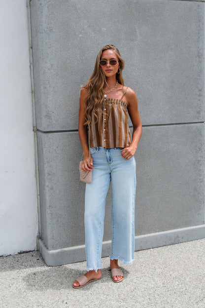 Woman in sunglasses wears the Sadie & Sage Striped Linen Cami - Camel, light blue jeans, and sandals by a concrete wall.