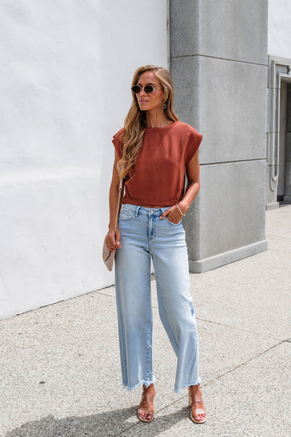 Woman in sunglasses, Sadie & Sage Ribbed Top - Rust, jeans, and sandals stands on sunny sidewalk by white and gray building.