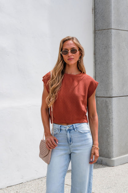 Woman in sunglasses wears the Sadie & Sage Ribbed Top - Rust, light jeans, and a textured bag by a white wall and gray pillar.