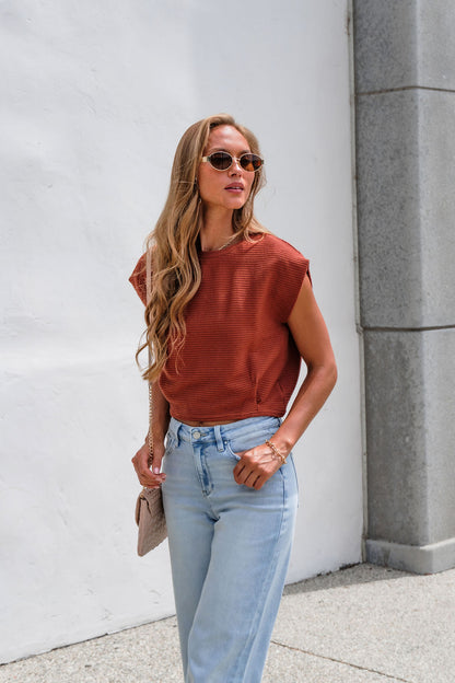 Woman with long hair in sunglasses wears Sadie & Sage Ribbed Top - Rust, light jeans, and a light handbag by a white wall.