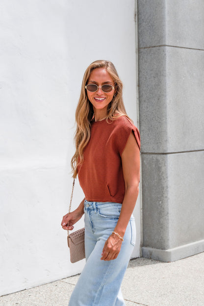 Woman in Sadie & Sage Ribbed Top - Rust, sunglasses, and light jeans smiles by a white wall, holding a purse.