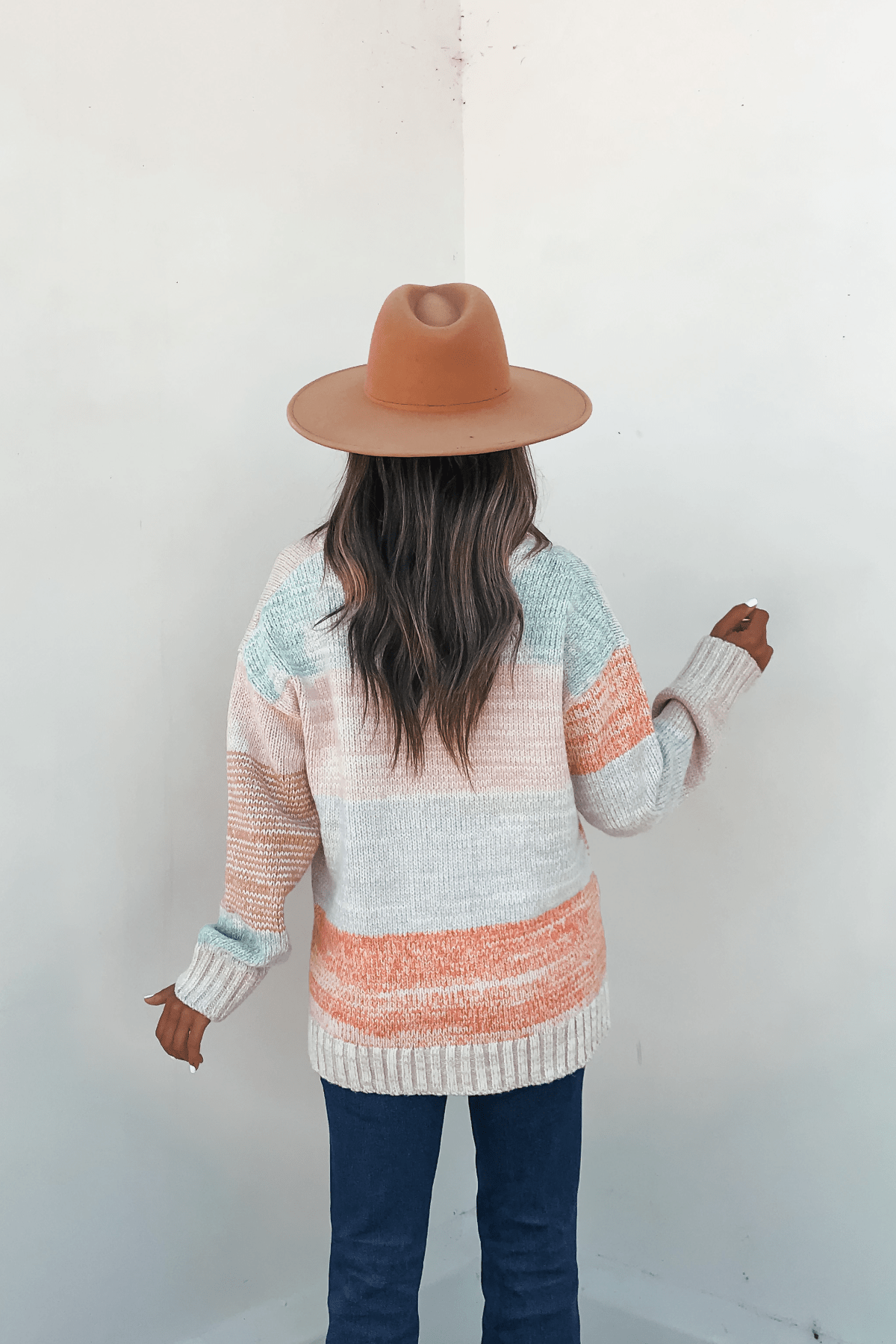 A woman in a tan hat and Oatmeal Color Block Button Down Cardigan stands with her back to the camera, facing a white wall.