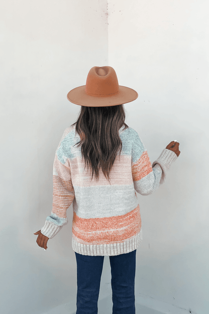 A woman in a tan hat and Oatmeal Color Block Button Down Cardigan stands with her back to the camera, facing a white wall.