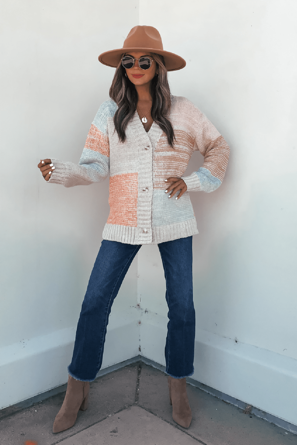 A woman in sunglasses and heeled boots wears the Oatmeal Color Block Button Down Cardigan and a wide-brim hat against a white wall.