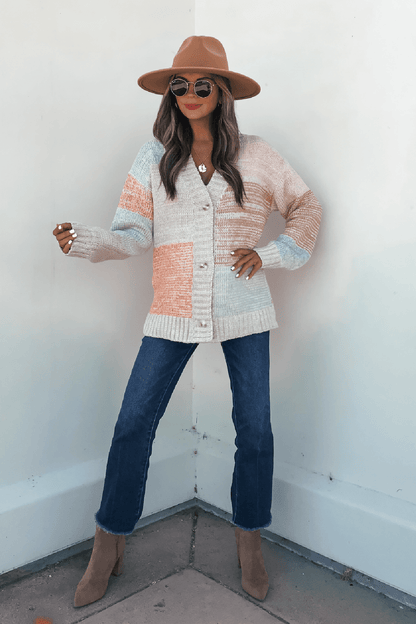 A woman in sunglasses and heeled boots wears the Oatmeal Color Block Button Down Cardigan and a wide-brim hat against a white wall.
