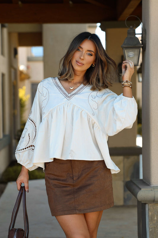 Woman with shoulder-length hair wears an Oatmeal Embroidered Babydoll Top and brown skirt, holding a bag outdoors.