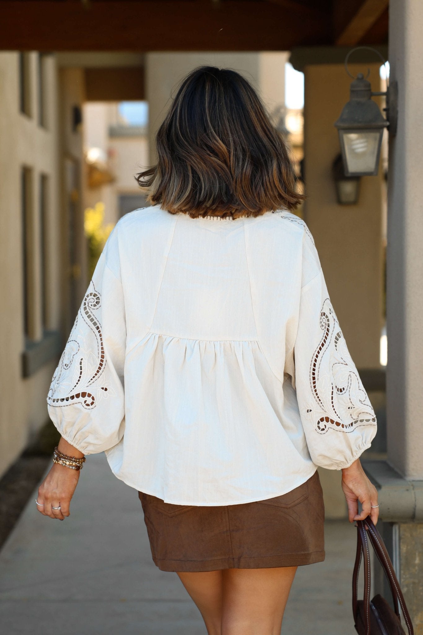 Woman with shoulder-length brown hair wears the Oatmeal Embroidered Babydoll Top and a brown skirt, walking outdoors.
